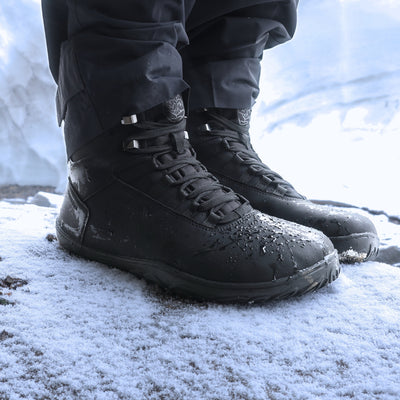 Käyttökuva noenmustista Uoma Talvi 3 -paljasjalkakengistä lumisella kivellä päivänvalossa. Kuvassa näkyy vedenpitävä pinta ja Vaistoa Pitoa™ -pohja.