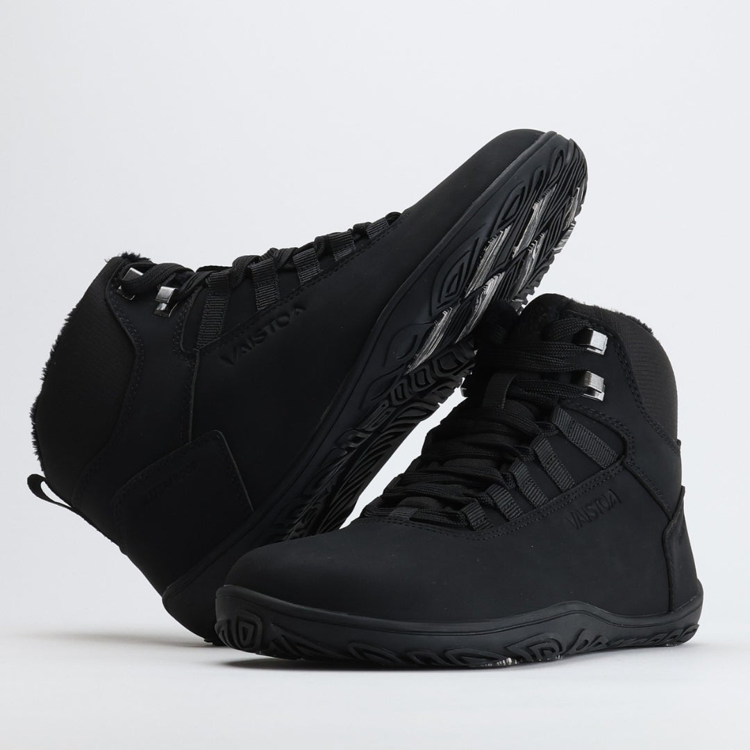 Noenmusta Vaistoa Uoma Talvi 3 -kenkäpari, jossa toinen kenkä nojaa toiseen ja näkyvissä on talvipohjan kitkapalat ja kuviointi.