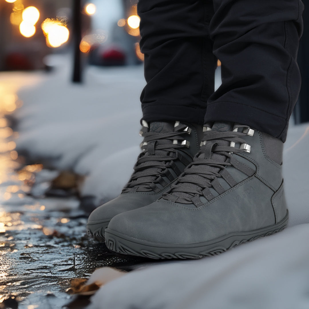 Käyttökuva pakkasharmaista Vaistoa Uoma Talvi 3 -paljasjalkakengistä iltavalaistulla kadulla. Lumi ja heijastuvat valot korostavat kengän urbaania tyyliä.