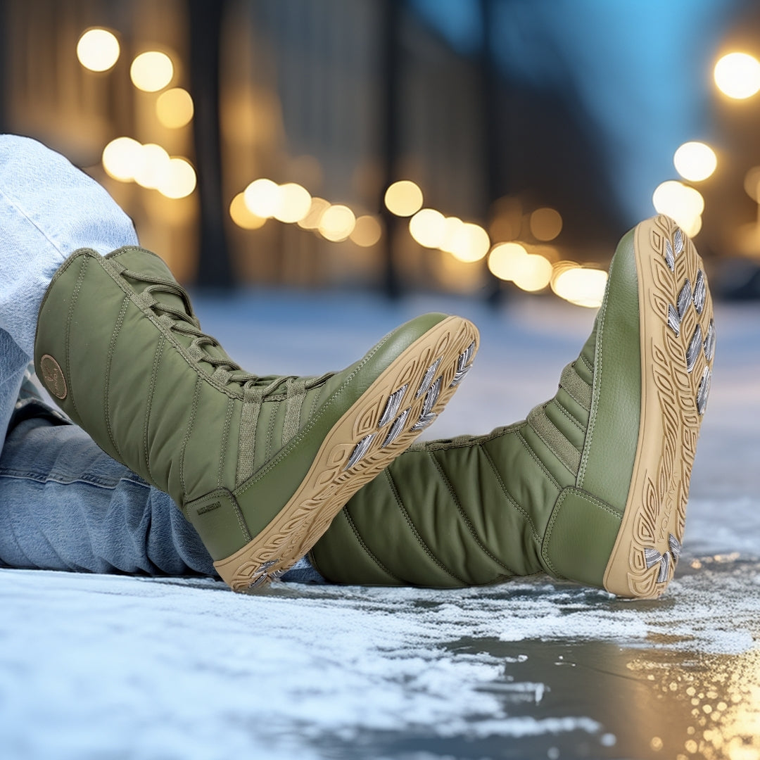 Vaistoa Hanki – korkeavartiset paljasjalkakengät talveen, väri oliivinvihreä. Käyttökuva: henkilö istuu lumella siniset farkut jalassa, taustalla kaupungin ilta ja valot luovat lämpimän talvitunnelman.