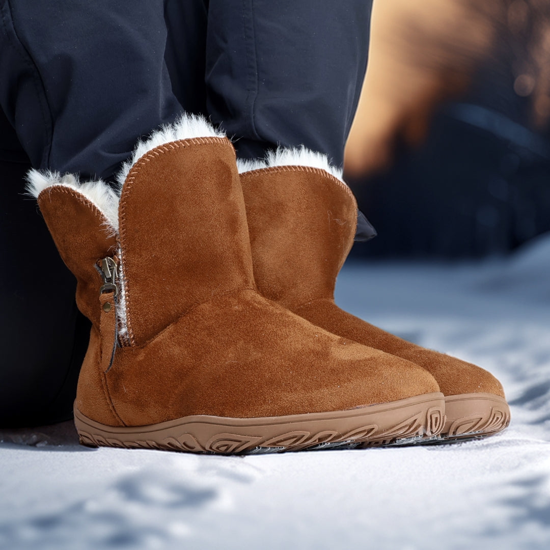 Vaistoa Huurre – paljasjalkabootsit talveen, väri karamellinruskea. Käyttökuva: etuviistosta kuvattuna, henkilö seisoo lumella Huurre-bootsit jalassa – kuva korostaa lämpöä, mukavuutta ja pitävää talvipohjaa.
