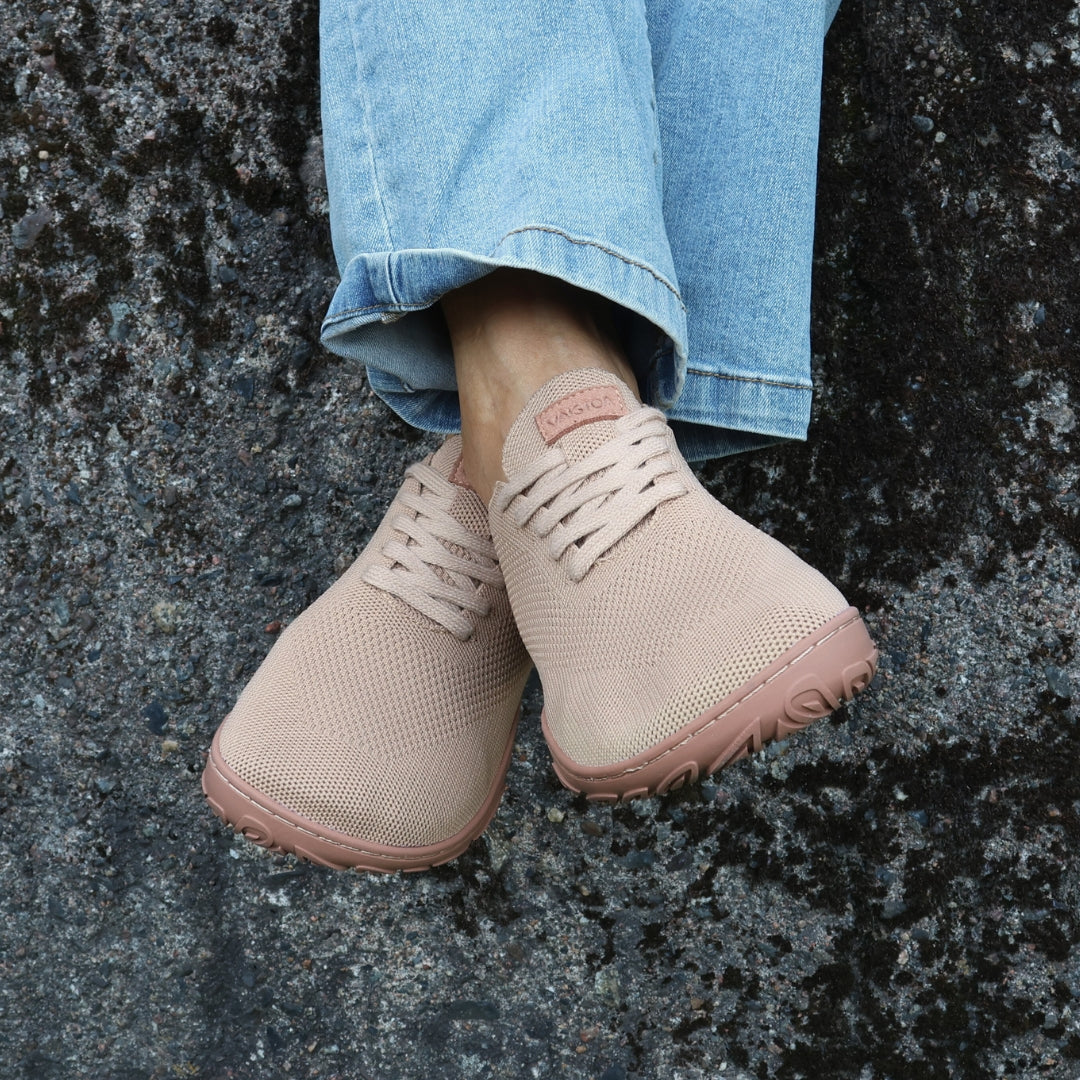 Vaistoa latte-väriset paljasjalkakengät sinisten farkkujen kanssa kiviseinää vasten istuen – minimalistiset ja leveävarvastilaiset kengät vapaa-aikaan.