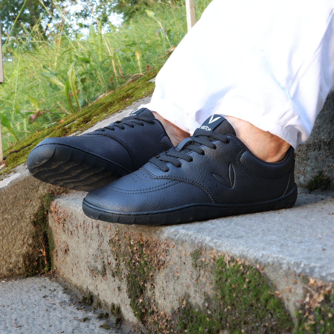 Vaistoa Katu 2 mustat paljasjalkasneakerit käytössä – henkilö istuu ulkona portailla kesäpäivänä valkoisissa farkuissa, kuvattuna läheltä.