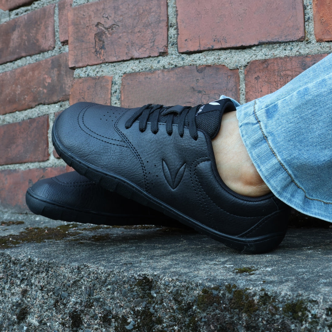 Vaistoa Katu 2 mustat paljasjalkasneakerit käytössä – henkilö istuu ulkona syksyisenä päivänä sinisissä farkuissa, jalat ilmassa tummapintaista seinää vasten.