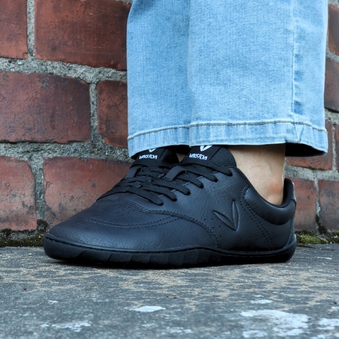 Vaistoa Katu 2 mustat paljasjalkasneakerit käytössä – henkilö istuu ulkona sinisissä farkuissa syksyisellä kivipinnalla, kuvattuna edestä.