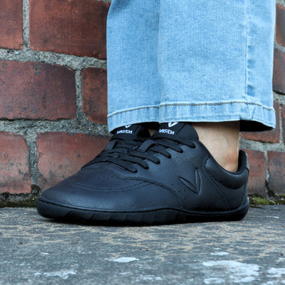 Vaistoa Katu 2 mustat paljasjalkasneakerit käytössä – henkilö istuu ulkona sinisissä farkuissa syksyisellä kivipinnalla, kuvattuna edestä.