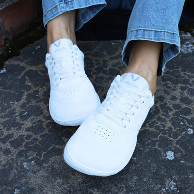 Vaistoa Katu 2 valkoiset paljasjalkasneakerit käytössä – henkilö istuu ulkona sinisissä farkuissa syksyisellä kivipinnalla, kuvattuna edestä.