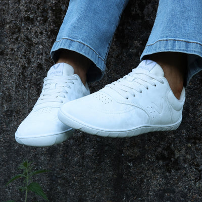 Vaistoa Katu 2 valkoiset paljasjalkasneakerit käytössä – henkilö istuu ulkona syksyisenä päivänä sinisissä farkuissa, jalat ilmassa tummapintaista seinää vasten.