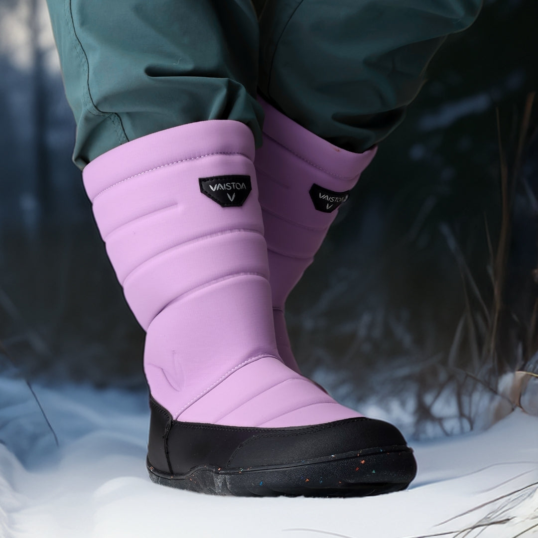 Vaistoa Kinos – lasten korkeavartiset paljasjalkakengät talveen, väri lila. Käyttökuva: etuviistosta kuvattuna sinisten housujen kanssa talviympäristössä.