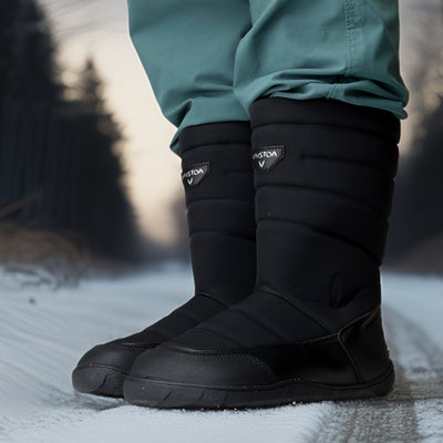 Vaistoa Kinos – lasten korkeavartiset paljasjalkakengät talveen, väri musta. Käyttökuva: etuviistosta kuvattuna lumisella tiellä sinisten housujen kanssa.