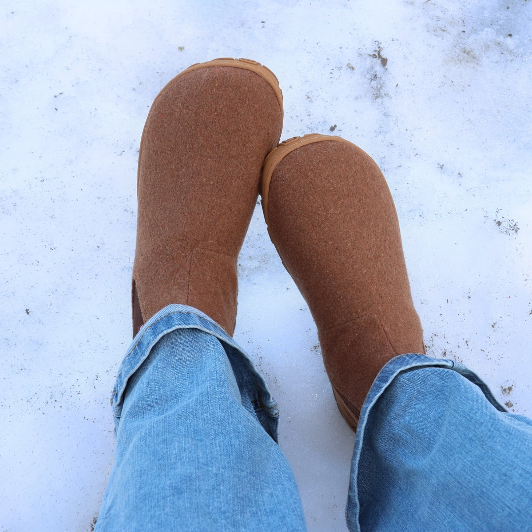 Ylhäältä otettu kuva henkilöstä, jolla on siniset farkut ja jalassa paahdetun mantelin väriset Vaistoa Sulo -huopanilkkurit. Lumi peittää maan ja kengät erottuvat selvästi, korostaen niiden leveää varvastilaa ja jalan luonnollista muotoa. Kuva välittää rauhallisen talvipäivän tunnelman.