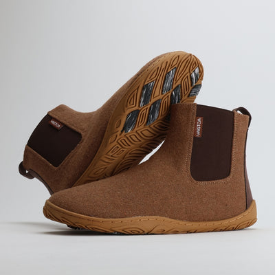 Kaksi Vaistoa Sulo -huopanilkkuria paahdetun mantelin sävyssä. Toinen kenkä nojaa toisen varteen, jolloin pohjan kitkapalat ja kengän sivuprofiili erottuvat selkeästi. Kuva tuo esiin minimalistisen ja luonnollisen muotoilun.