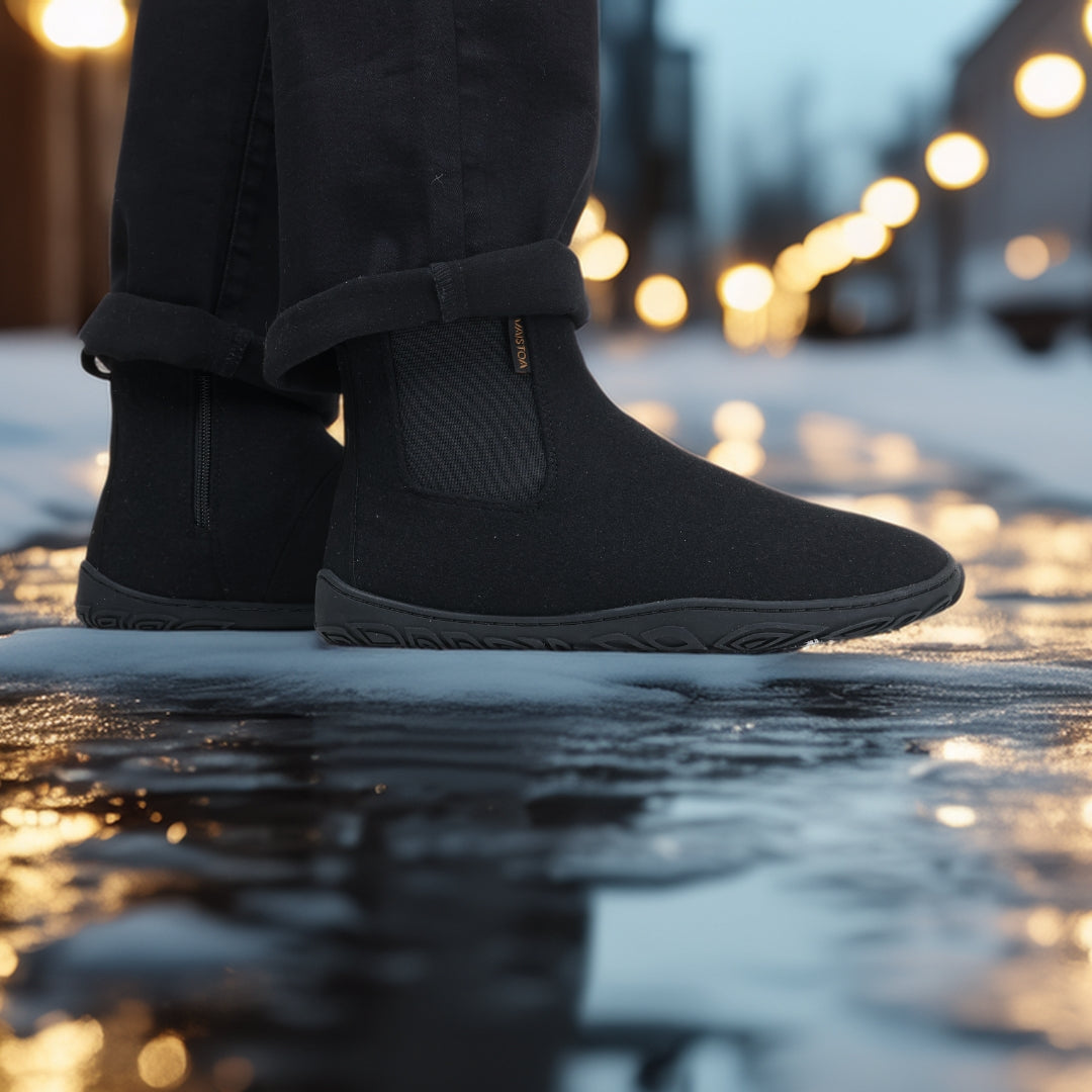 Käyttökuva, jossa henkilö seisoo samettisen mustat Vaistoa Sulo -huopanilkkurit jalassa valaistulla, kostealla talvikadulla. Kuvakulma etuviistosta korostaa kengän tyylikästä minimalistisuutta ja pitävää pohjarakennetta.