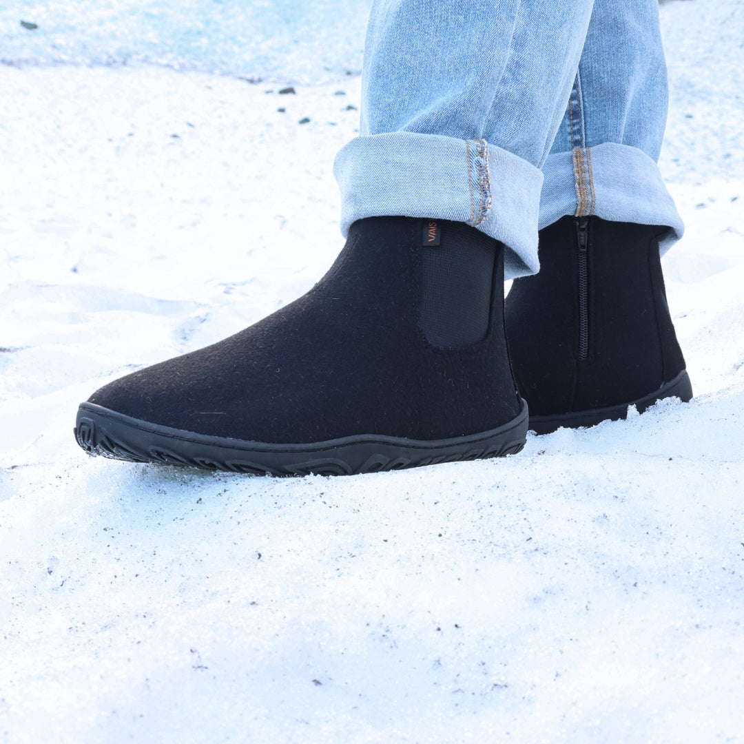 Käyttökuva, jossa henkilö seisoo lumella sinisissä farkuissa ja samettisen mustissa Vaistoa Sulo -huopanilkkureissa. Kuvakulma etuviistosta korostaa kengän pehmeää huopapintaa ja mukautuvaa pohjarakennetta.